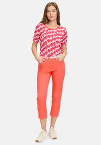 Un haut à manches courtes et col rond présente un motif géométrique rose. Associé à un pantalon court corail et des baskets légères. Une ceinture blanche ajoute du contraste.