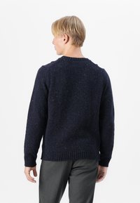 Granatowy sweter dzianinowy z teksturowanym, nakrapianym wzorem, z okrągłym dekoltem oraz prążkowanymi mankietami i dołem. Nosi się go na szarych spodniach.
