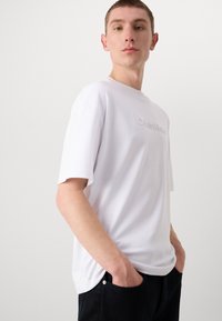 White short-sleeve T-shirt with a crew neckline, featuring a subtle embossed logo on the front and a smooth, lightweight fabric.