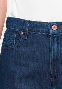 Close-up van de taille van donkerblauwe denim jeans met koperen klinknagels, riemlussen en een wit geribbeld shirt dat erin is gestopt.