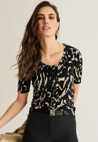 Vrouw met lange golvende bruine haren, gekleed in gouden creolen, een zwart en beige top met dierenprint, en zwarte broek met een riem met een "M" gesp.