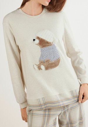 Pull long gris clair avec un ours brun portant un pull en tricot bleu et un bonnet tricoté blanc. Ourlet inférieur à motifs à carreaux et pantalon.