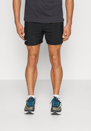 Homme portant un short de sport noir, des baskets noires et bleues, et un t-shirt gris foncé, debout devant un fond uni.