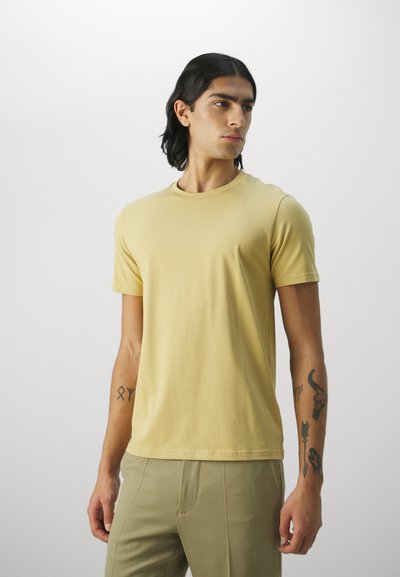 Camiseta de manga corta lisa en algodón amarillo claro, con cuello redondo y diseño ajustado. Llevada con pantalones ligeros verdes a medida, mostrando tatuajes visibles.