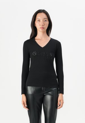 Black long-sleeve V-neck t-shirt with rhinestone "Guess est. 1981" logo at the chest. Fitted design and soft fabric texture.