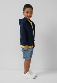 Sudadera de cremallera navy, camiseta amarilla, pantalones cortos de denim y zapatillas blancas. La sudadera tiene un bolsillo frontal, un corte relajado y puños acanalados.