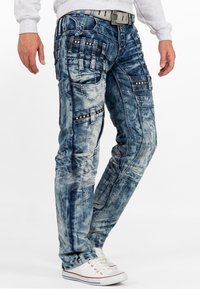 Pantaloni di jeans azzurri chiari con molteplici tasche, cuciture decorative e una finitura strutturata. Gli accenti includono accessori neri e argentati.