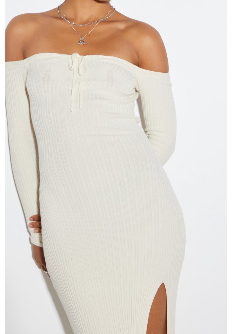 Femme portant une robe en maille côtelée blanche à épaules dénudées, manches longues et fente avant, accessoirisée de colliers argentés superposés.