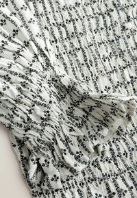 Material alb cu dungi florale negre, cu smocuri adunate și detalii cu volane la mâneci.