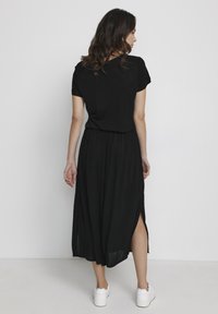 Abito maxi nero con una texture morbida, maniche corte e spacco laterale. Indossato con sneaker bianche, che mostrano una vestibilità rilassata e un design fluido.