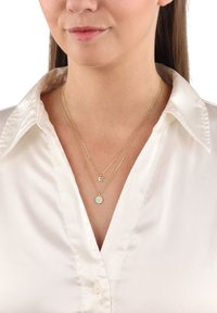 Guldpläterade lagerhalsband med en stjärncharms och en rund berlock med en strukturerad yta, bärs över en vit skjorta med knappar.