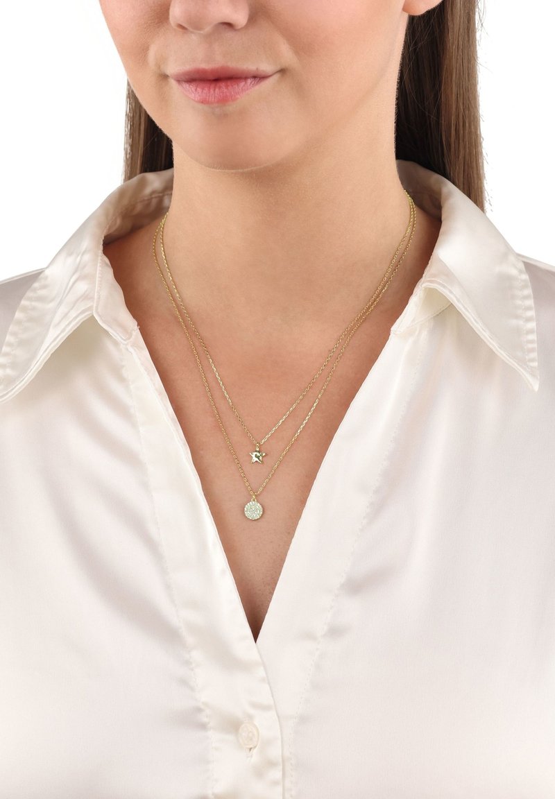 Guldpläterade lagerhalsband med en stjärncharms och en rund berlock med en strukturerad yta, bärs över en vit skjorta med knappar.