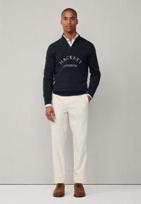 Navy-blaues gestricktes Sweater mit "HACKETT LONDON" Logo, weißem Hemdkragen, cremefarbener Hose und braunen Lederschuhen. Das Modell steht aufrecht vor einem schlichten Hintergrund.