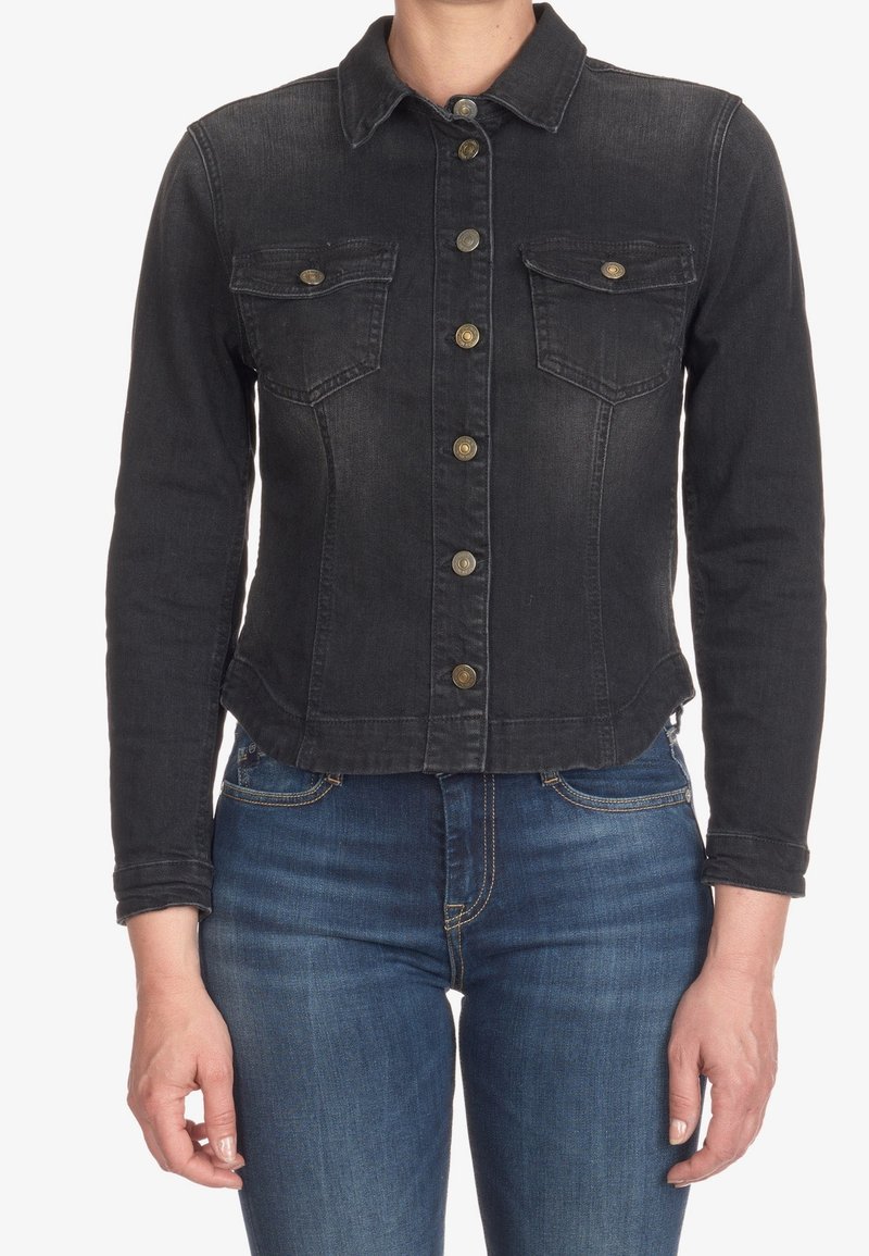 Kvinna iklädd en figurnära svart denimjacka med knäppning och bröstfickor, tillsammans med mörkblå jeans.