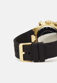 Correa de reloj de goma negra con hebilla y herrajes metálicos dorados. Presenta un reverso texturizado y la carátula del reloj redonda visible en el fondo.