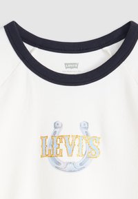 Hvid skjorte med en marineblå halsudskæring. Har et blåt hestesko-grafik og ordet "LEVI'S" i guldglitter. Blødt bomuldsblandingsmateriale.