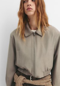 Khaki zip-up jakke med kollaret hals, elastisk talje og forlommer. Glat stof med subtile teksturdetaljer.