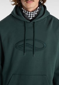 Felpa con cappuccio verde scuro caratterizzata da un logo in rilievo, cordini e una tasca frontale. Tessuto morbido con una consistenza liscia, ideale per essere indossata a strati.