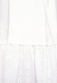 Vêtement blanc présentant un tissu supérieur lisse et une partie inférieure froncée avec une broderie florale en œillets et de petits détails découpés.