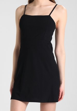 Mujer con un minivestido negro sin mangas y tirantes finos sobre un fondo claro y liso.