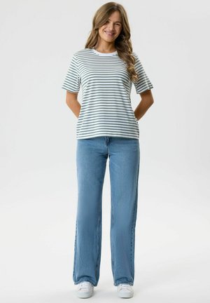 Mlada žena se smiješi noseći bijelu majicu s zelenim prugama, svijetlo plave traperice i bijele tenisice, stojeći s rukama iza leđa.