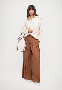 Cream-colored shirt with textured cut-out design, paired with wide brown trousers. Accented with a structured beige bag and beige shoes.