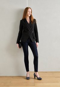Černý blazer s předním zavinovacím designem a stříbrným kroužkovým detailem, spárovaný s tmavými úzkými džíny a šedými vysokými podpatky, držící malou černou kabelkou.