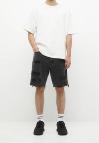 T-shirt en coton blanc surdimensionné associé à un short en denim noir usé avec de multiples déchirures, des bords effilochés et des baskets noires.