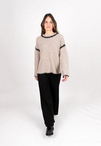 Beige gestrickter Pullover mit schwarzem Zickzack-Muster am Kragen und an den Ärmeln, kombiniert mit schwarzen weit geschnittenen Hosen und schwarzen Stiefeletten.