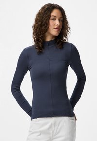 Jovem mulher com cabelo encaracolado veste uma blusa de manga longa listrada azul-marinho com colarinho alto e calças de tom claro, posando contra um fundo simples.