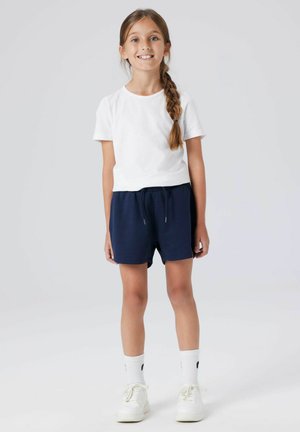 Niña joven con el cabello largo trenzado, vestida con una camiseta blanca, pantalones cortos azul marino, calcetas blancas y zapatillas blancas, de pie frente a un fondo liso.