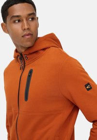 Orange zip-hættejakke i blødt stof; har en sort lynlåslomme på brystet og logo på ærmet; tætsiddende design med hætte.