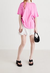 Světlý růžový oversized bavlněný tričko, bílé volánkové sukně, černá strukturovaná kabelka a robustní černé sandály s křížovými pásky.