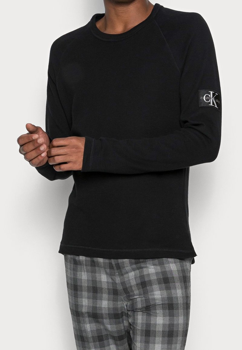 Man som bär en svart långärmad skjorta från Calvin Klein och grå rutiga byxor, med händerna mot bröstet framför en enhetlig bakgrund.