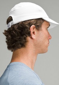 Witte cap met een gebogen rand en een gestructureerd oppervlak, met een verstelbare riem aan de achterkant. Model weergegeven vanaf de zijkant.