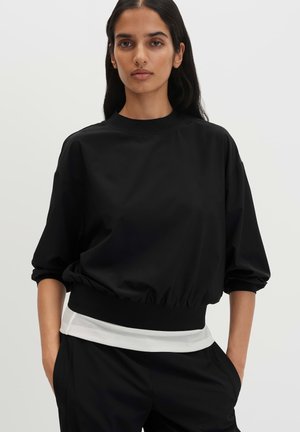 Femme aux longs cheveux foncés portant un haut noir ample avec les manches retroussées et un pantalon noir, les mains dans les poches, sur fond blanc.