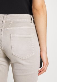 Beiga denimjeans med medelhög midja, bakfickans stygn och en slät textur. Hand som vilar på sidan, neutral bakgrund.