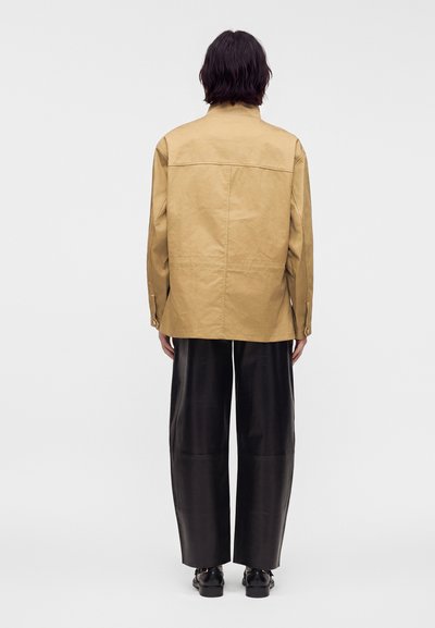 Personne aux cheveux foncés mi-longs, portant une veste beige, un pantalon large noir et des chaussures noires, debout, le dos tourné vers l’avant.
