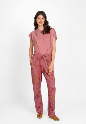 Vrouw met donker haar, een roze gestreept shirt en roze broek met bloemmotief, staand met één hand in haar zak en glimlachend.