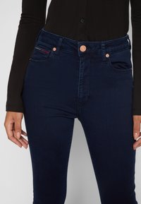 Pantalones vaqueros ajustados de color azul oscuro, hechos de denim, con herrajes de cobre, bolsillos frontales y sutiles detalles de costura, combinados con una blusa negra de manga larga.