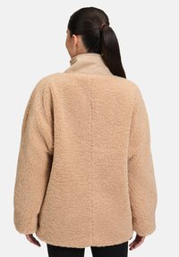 Veste en fausse fourrure beige avec une texture douce, coupe oversize et col haut. Présente une structure de couture simple et des manches larges.
