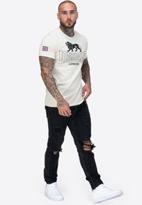 Krem majica kratkih rukava s crnim grafičkim motivom lava i natpisom "LONSDALE LONDON", uparena s istrošenim crnim trapericama i bijelim tenisicama.