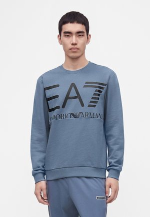 Młody mężczyzna z krótkimi czarnymi włosami, ubrany w niebieską bluzę EA7 Emporio Armani oraz pasujące niebieskie spodnie, stoi na tle jednolitego białego tła.