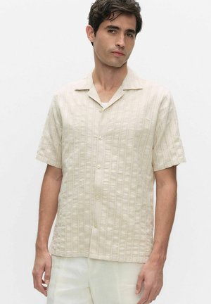 Man draagt een beige, gestructureerd overhemd met korte mouwen en kraag, gecombineerd met een lichtgekleurd broek, tegen een effen achtergrond.