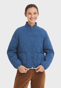 Veste matelassée bleue avec fermeture éclair sur le devant, col haut et deux poches. Présente des coutures courbées et une coupe décontractée. Matériau texturé de poids moyen.