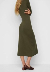 Femme portant une robe longue olive à manches longues et des ballerines marron, vue de profil sur un fond blanc uni.