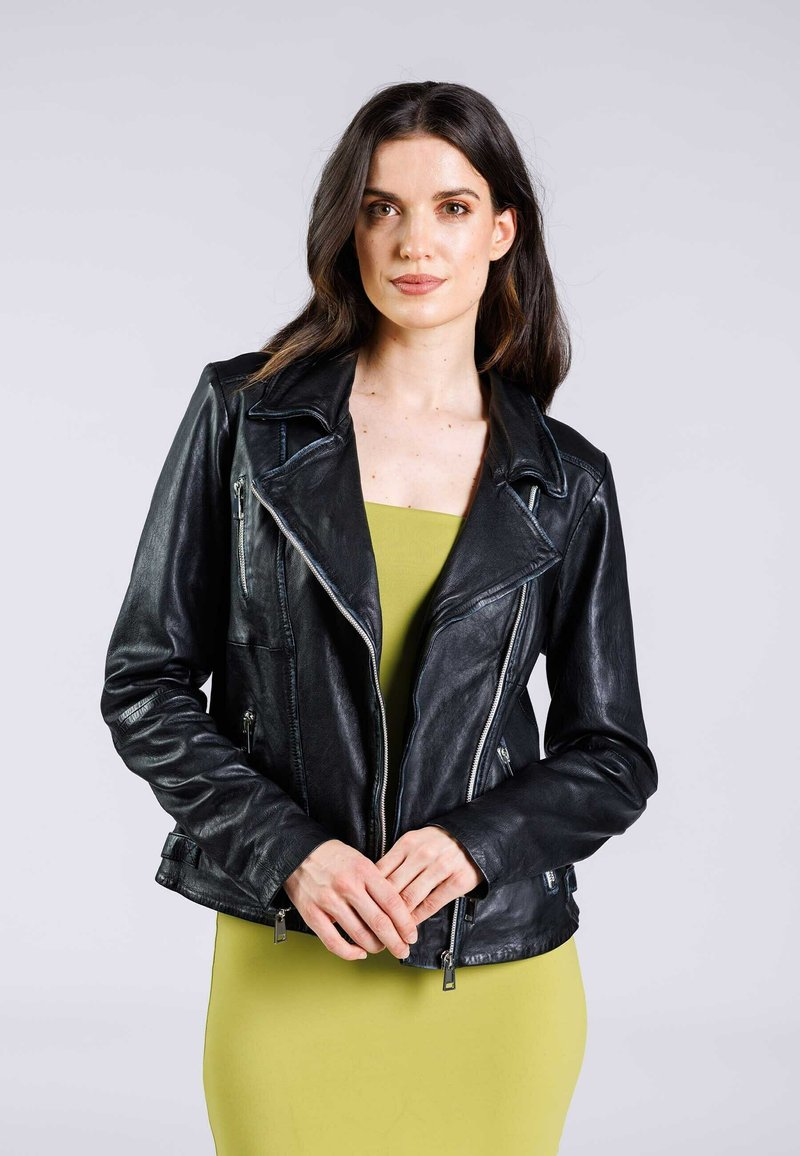 Giacca da motociclista in pelle nera con colletto a punte, dettagli con zip e design aderente, abbinata a un vestito verde. Texture liscia.