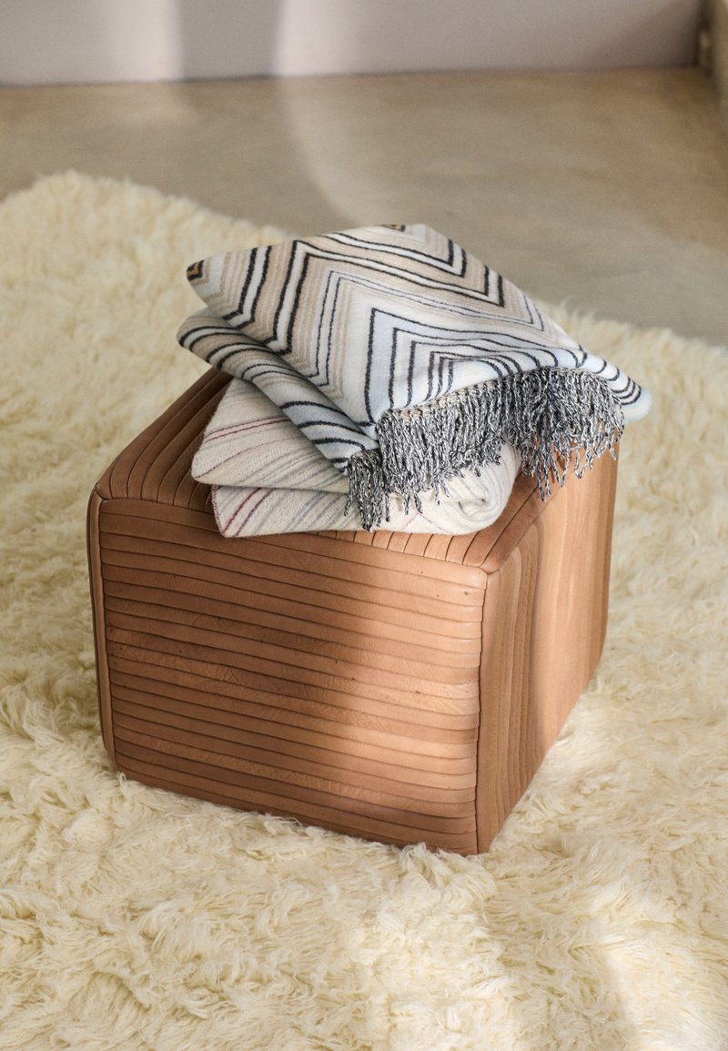 Pohjakuutioapupöytä puusta, jossa on pystysuorat urat, päällä kolme taitettua vilttiä valkoisissa ja mustissa sahalaitakuviolla, asetettuna pörröiselle matolle.