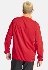 Rote Sportjacke mit orangefarbenen Akzenten, glatte Textur, lange Ärmel, hoher Kragen, mit elastischen Bündchen und einem lässigen Schnitt.