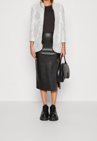 Kobieta ubrana w srebrną marynarkę z cekinami, czarną bluzkę, czarną skórzaną spódnicę midi z rozcięciem, masywne czarne sneakersy i trzymająca czarną torebkę.